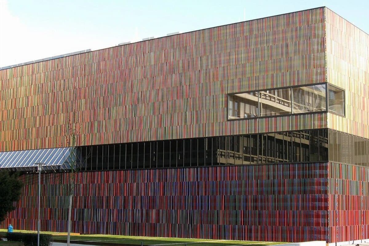 A museum building covered in thin vertical rods in red, orange, green and blue, with a band of dark glass above a covered walkway
